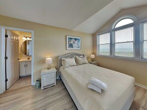 Un dormitorio con cama, mesitas de noche, un armario auxiliar, una ventana con persianas, una puerta que da a un baño y un cuadro en la pared.