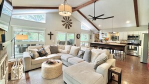 A spacious living room with a sectional sofa, a round ottoman, and a dining area with a bar. - Honey Haus On The Horseshoe - Guadalupe River + Listen to Whitewater Amphitheater Concerts Out Back! (Canyon Lake)