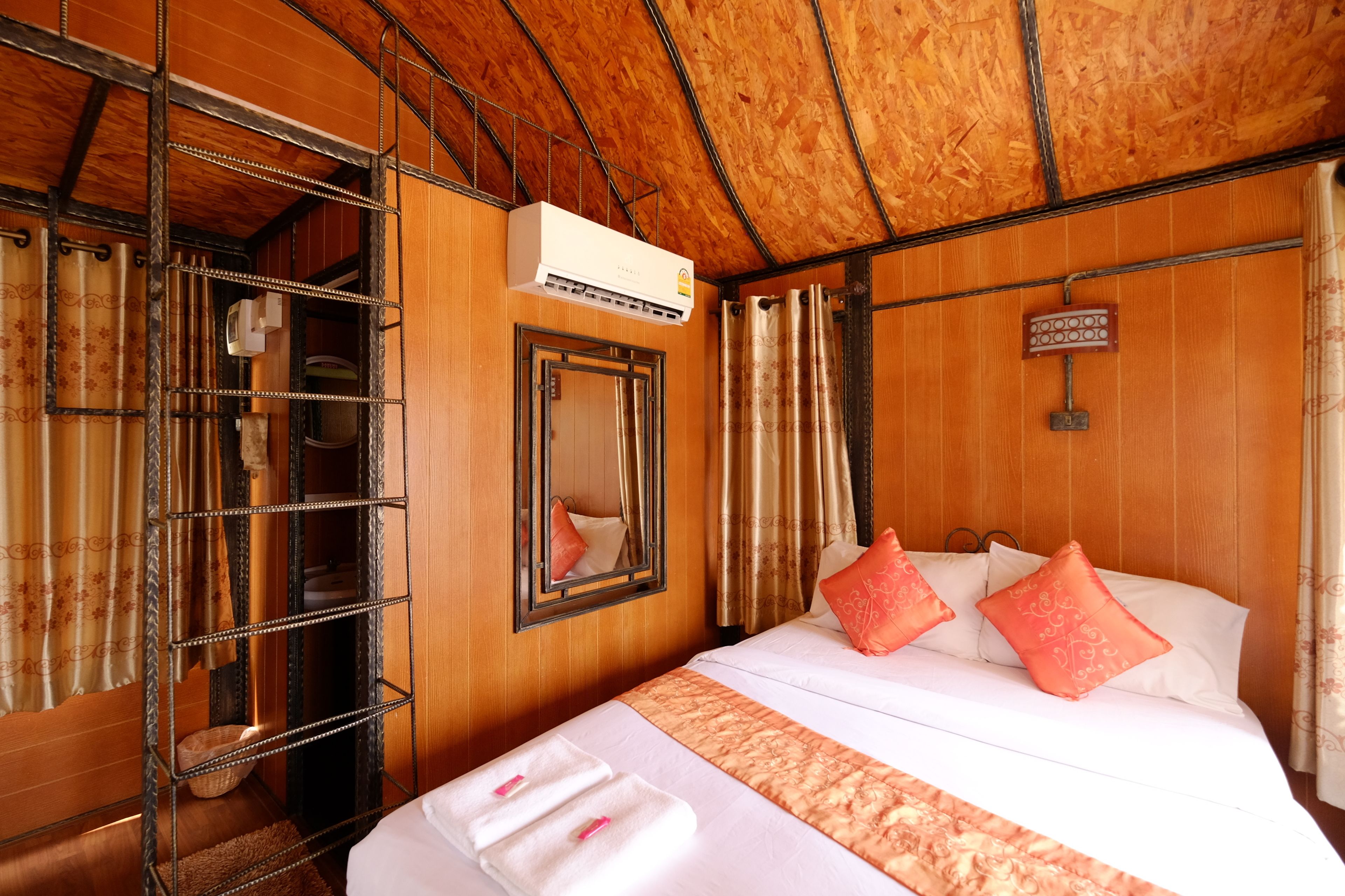 A hotel room with a wooden ceiling, a bed with white bedding and pink pillows, a metal shelf unit, and an air conditioning unit.