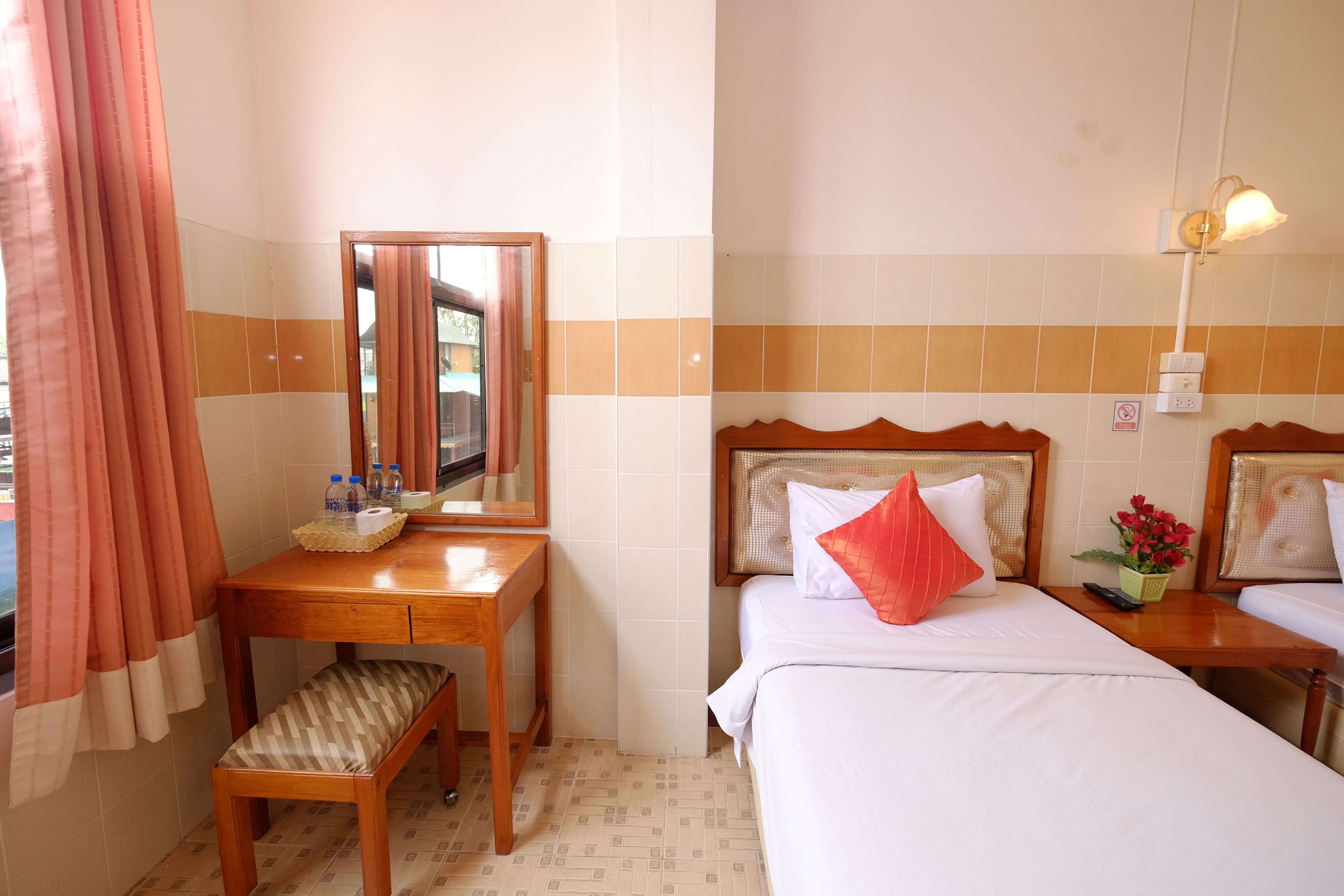 A hotel room with a wooden desk, a bench, a bed with white bedding and a red pillow, a bedside table with a remote control and a flower vase, and a mirror on the wall.