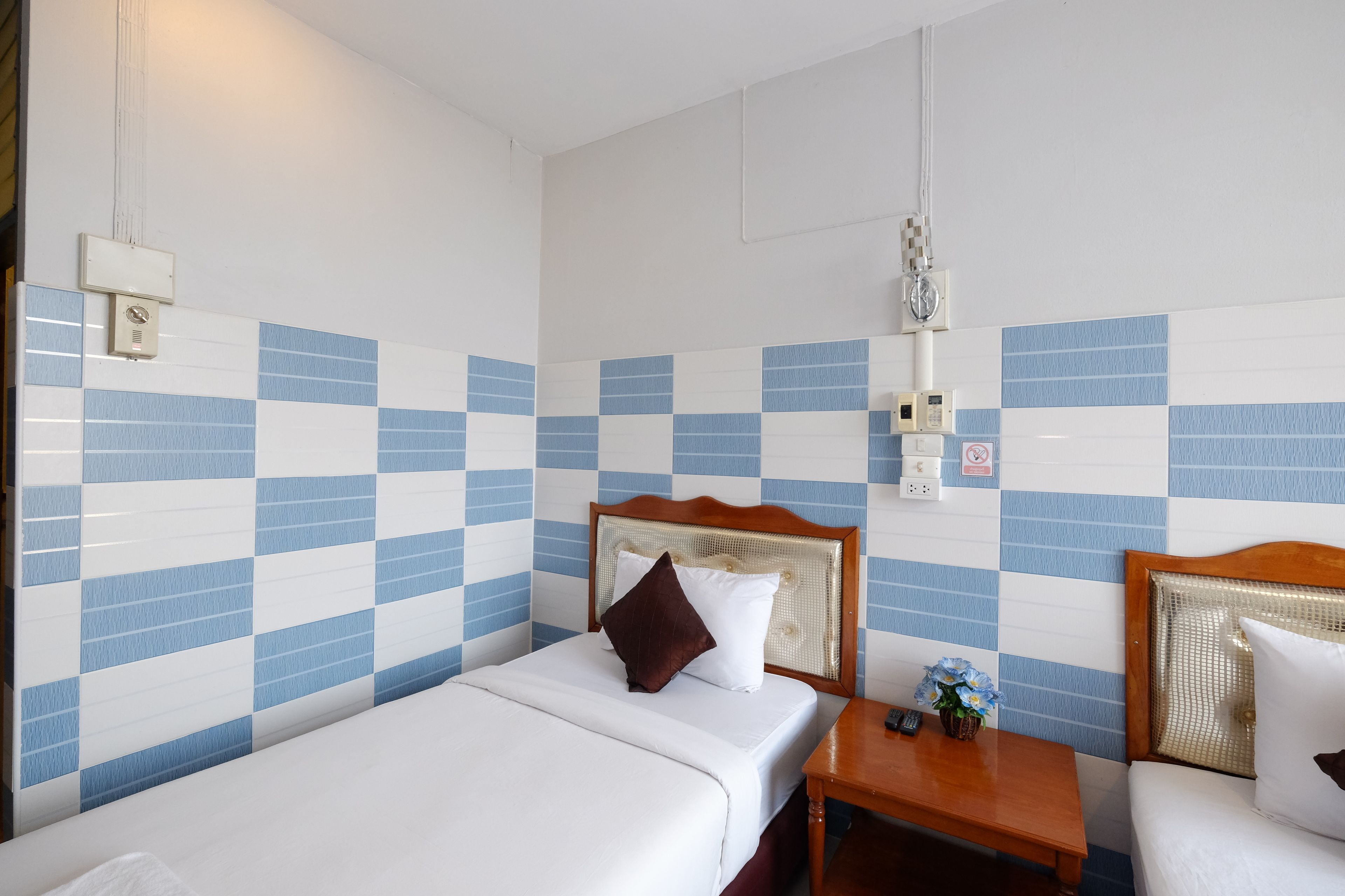 Habitación de hotel con pared de baldosas azules y blancas a cuadros, cabecera de madera, mesita de noche con control remoto y una pequeña planta, y cama con ropa blanca y almohada marrón.