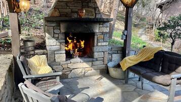 An outdoor stone fireplace with a fire, surrounded by wooden furniture and a yellow blanket.