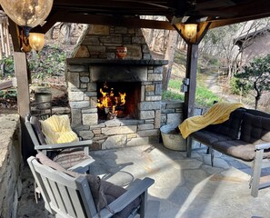 An outdoor stone fireplace with a fire, surrounded by wooden furniture and a yellow blanket. - Hot tub, Game room, Rustic elegance, Fresh eggs, Private, 10 miles to AVL. (Fairview)