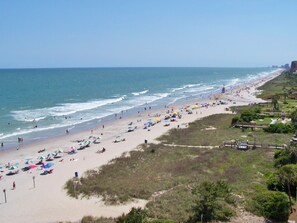 A beach with umbrellas and people, a boardwalk, and buildings in the background.