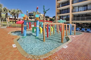 A water play area with colorful structures and a pool, surrounded by a brick walkway and palm trees.