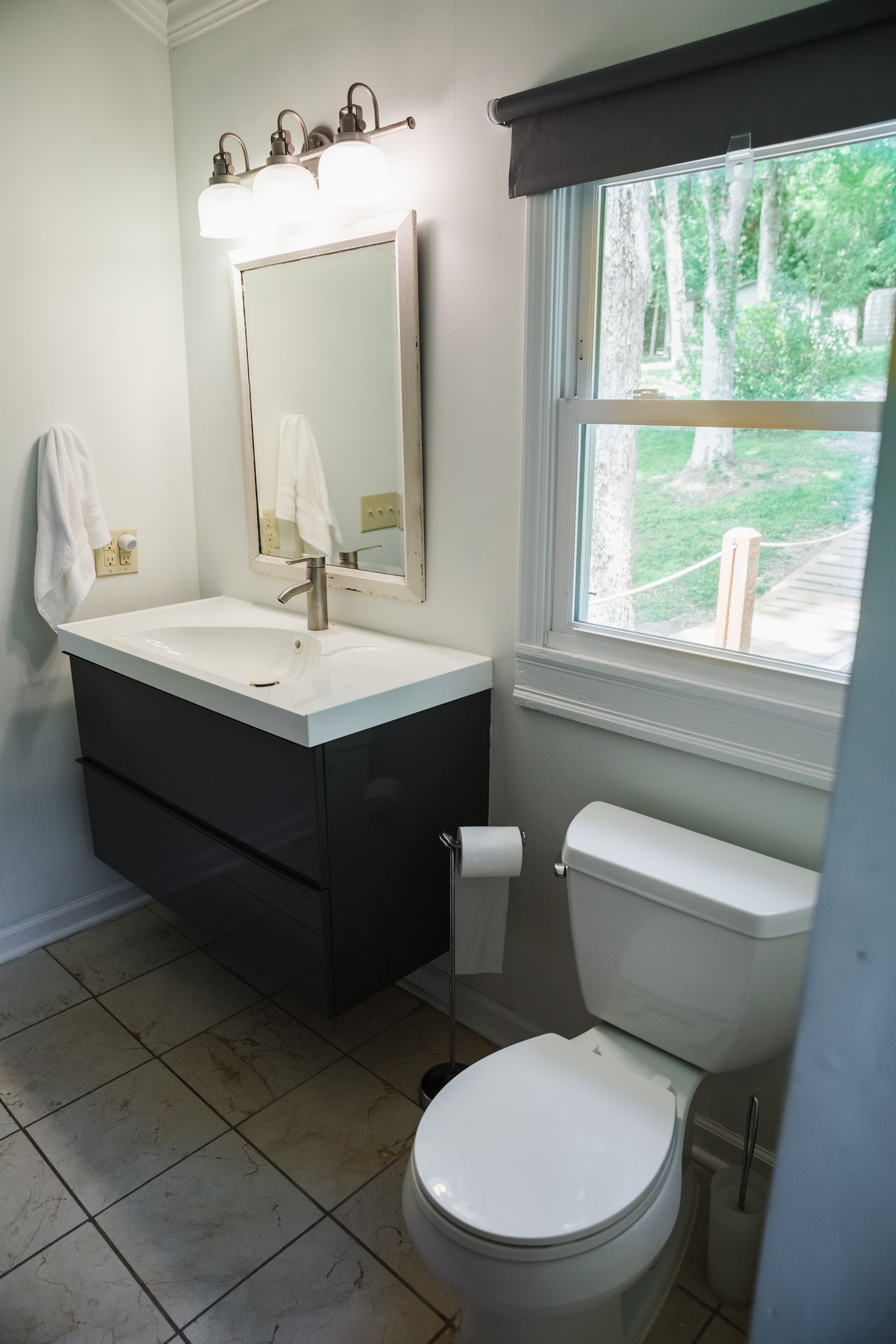 A bathroom with a white sink, dark vanity, a toilet, and a window with a view of trees.