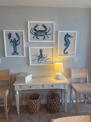 A room with a white desk, wicker chairs, a lamp, and framed seahorse and octopus artwork on the wall.