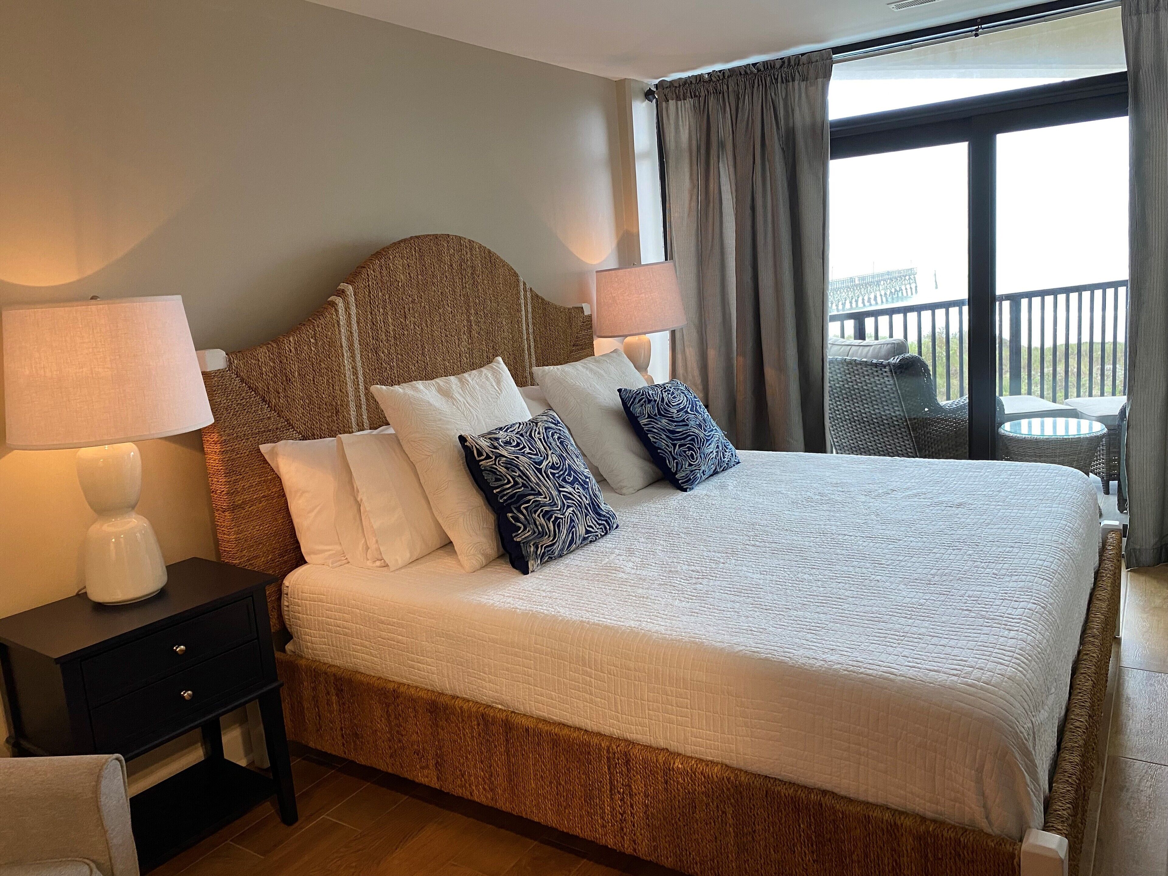 A hotel room with a large bed, two bedside tables with lamps, a view of the balcony, and a wicker chair.