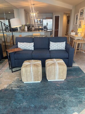 A modern living room with a dark blue sofa, two matching ottomans, a glass coffee table, and a kitchen in the background.