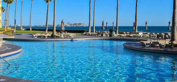 A pool area with lounge chairs and palm trees, a clear view of the ocean, and a distant island.