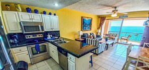 A kitchen with white cabinets, a microwave, and a stove. There is a dining area with striped sofas and a view of the ocean.