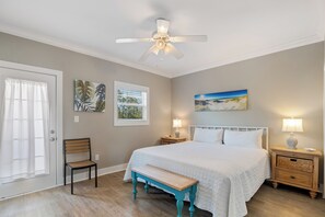 Une chambre à coucher comprenant un lit, un ventilateur de plafond, un banc, une chaise, une table de chevet avec une lampe et des œuvres d’art accrochées aux murs.