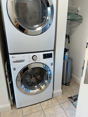 Une machine à laver et un sèche-linge superposés dans une pièce dont le sol est recouvert de carreaux et où se trouve une poubelle en métal.