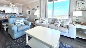 A modern living room with a sofa, armchair, and coffee table, a kitchen area, and a large window with curtains.