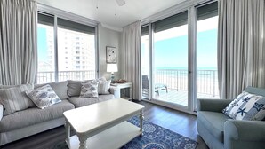 A spacious living room with a sofa, a coffee table, and a chair. There is a balcony with a view of the ocean and buildings.