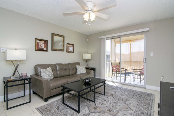 A living room with a brown leather sofa, a glass coffee table, a black side table, and a ceiling fan. There are two wall-mounted lamps and a large window with a view of a patio area.