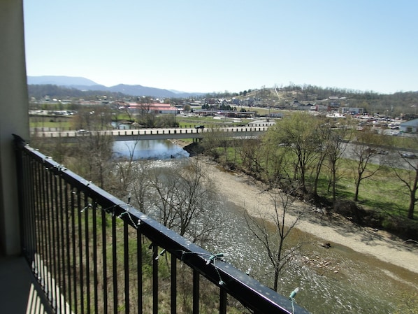 A view from a balcony overlooking a river, a bridge, and a town with buildings and mountains in the distance. - 50.00 October-stay gift card! Luxurious Condo, Riverfront w/ Mountain Views! (Sevierville)