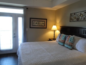 A hotel room with a bed, a nightstand with a lamp, a framed artwork on the wall, and a window with blinds.