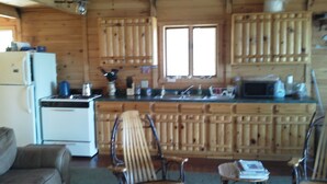 A cabin-style kitchen with wooden cabinets, a refrigerator, and a stove. There is a window providing natural light and a seating area with a couch and chairs.