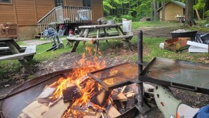 A rustic outdoor setting with a fire pit, picnic tables, and a wooden cabin in the background.