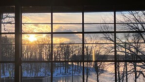Vista a través de una ventana con diseño de cuadrícula, mostrando un paisaje nevado, árboles y una masa de agua al atardecer.