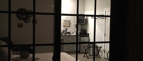 A room with a bed, a small table, and a chair. There is a kitchen area visible through a partition.