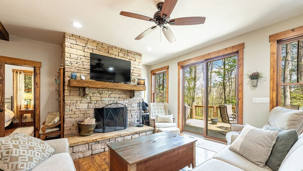 A cozy living room with a stone fireplace, comfortable seating, and large windows offering a view of the outdoors.