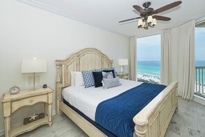 Quarto com cama, mesa de cabeceira, relógio, ventilador de teto e vista para o oceano.