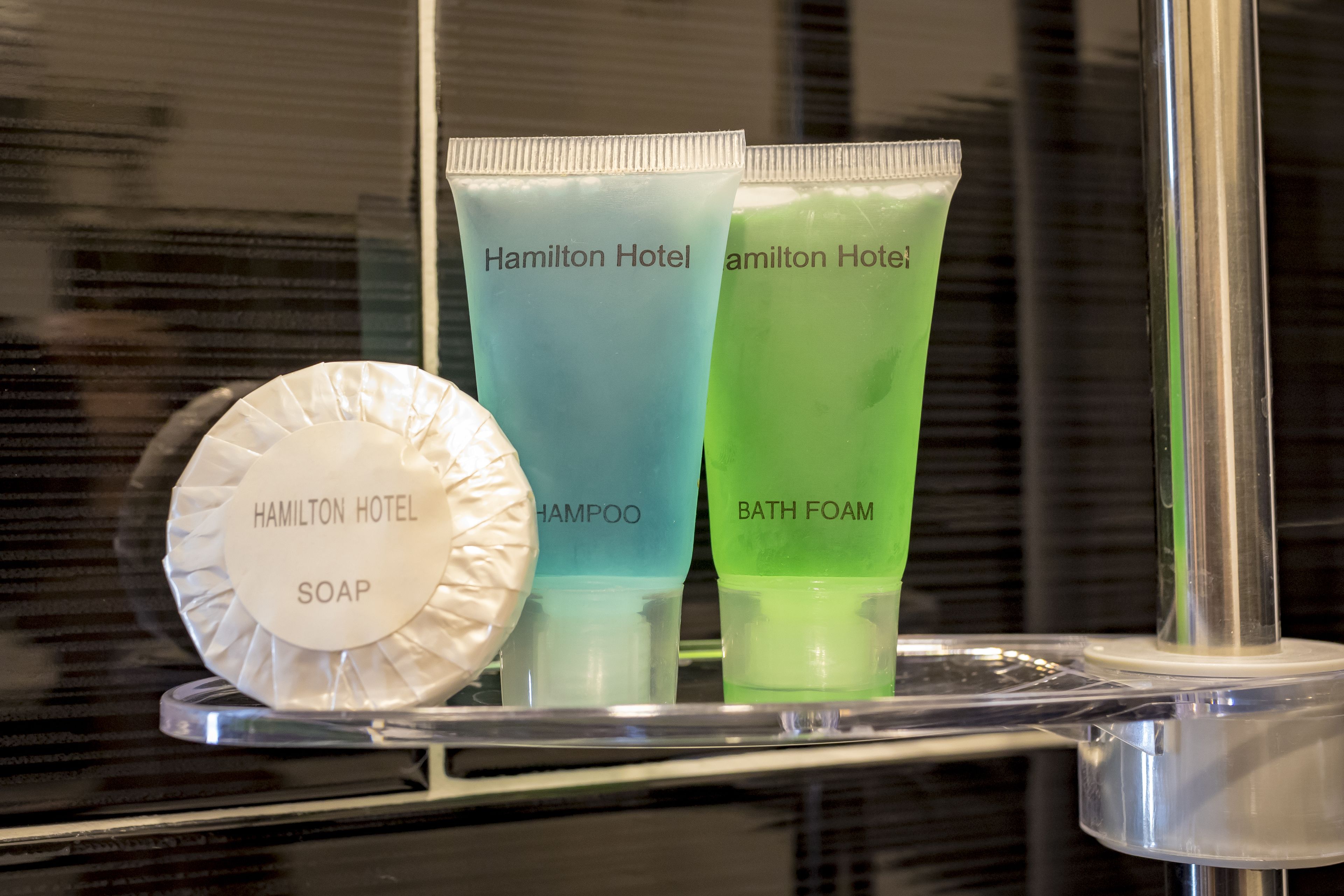 A bathroom shelf with toiletries labeled 'Hamilton Hotel' including soap, shampoo, and bath foam.