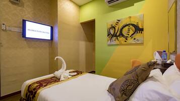 A hotel room with a bed, a wall-mounted TV displaying "HAMILTON HOTEL", an air conditioner, a painting, and a telephone.