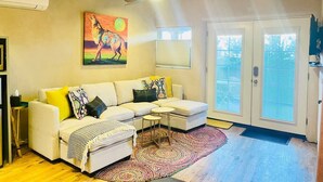 A living room with a beige sectional sofa, a small coffee table, and a colorful rug. There is a painting on the wall and a door leading outside.