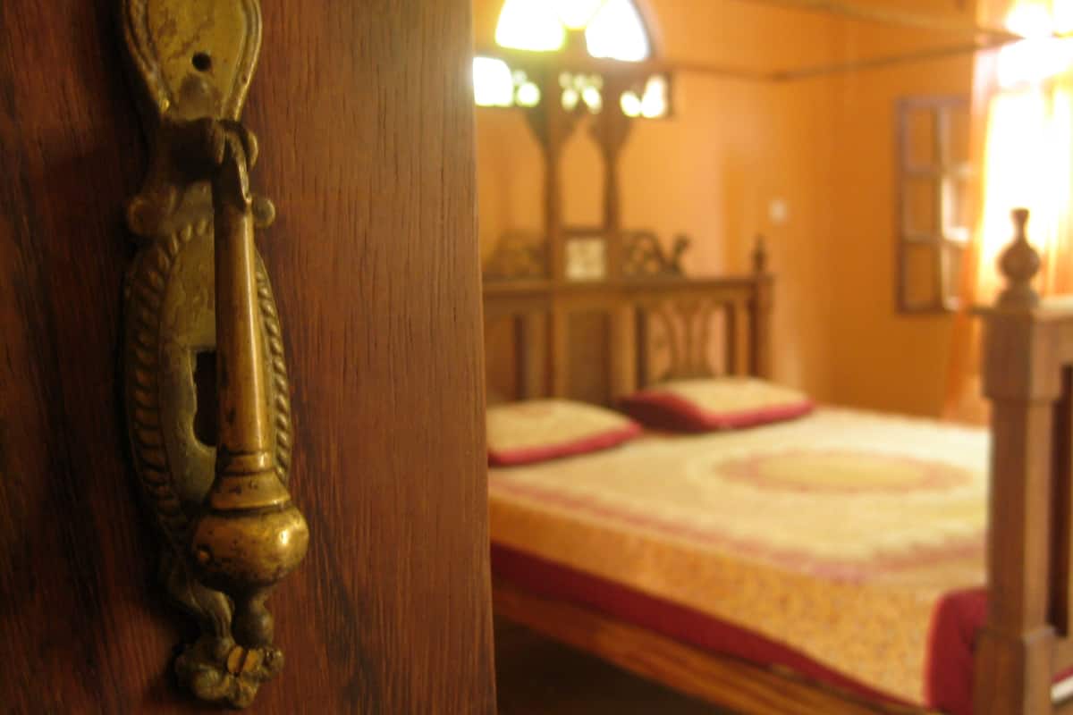 Una puerta de madera con un aldabón de bronce y una cama con colcha roja y blanca al fondo.
