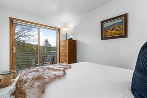 A bedroom with a large bed, a wooden dresser, a painting on the wall, and a view through a sliding glass door.