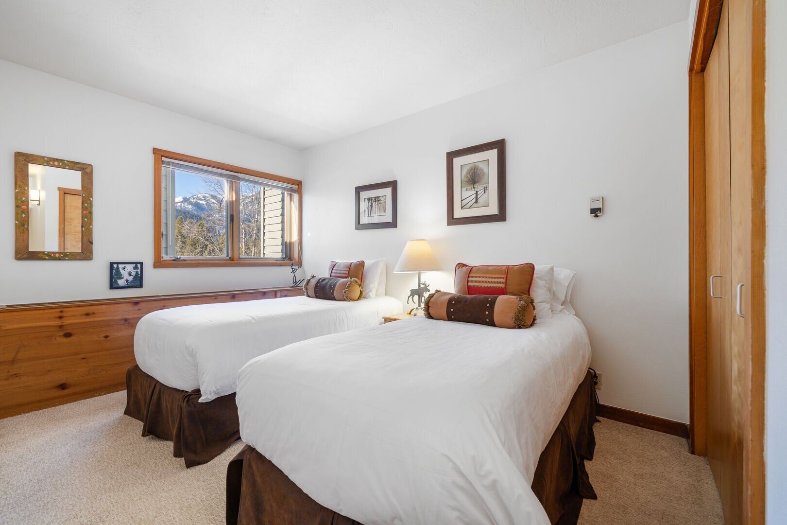 A bedroom with two beds, a window, a lamp, and framed pictures on the wall.