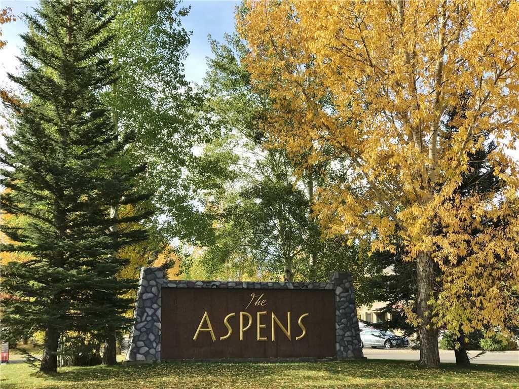 A sign for 'The ASPENS' with a stone wall and a large tree with yellow leaves in the background.