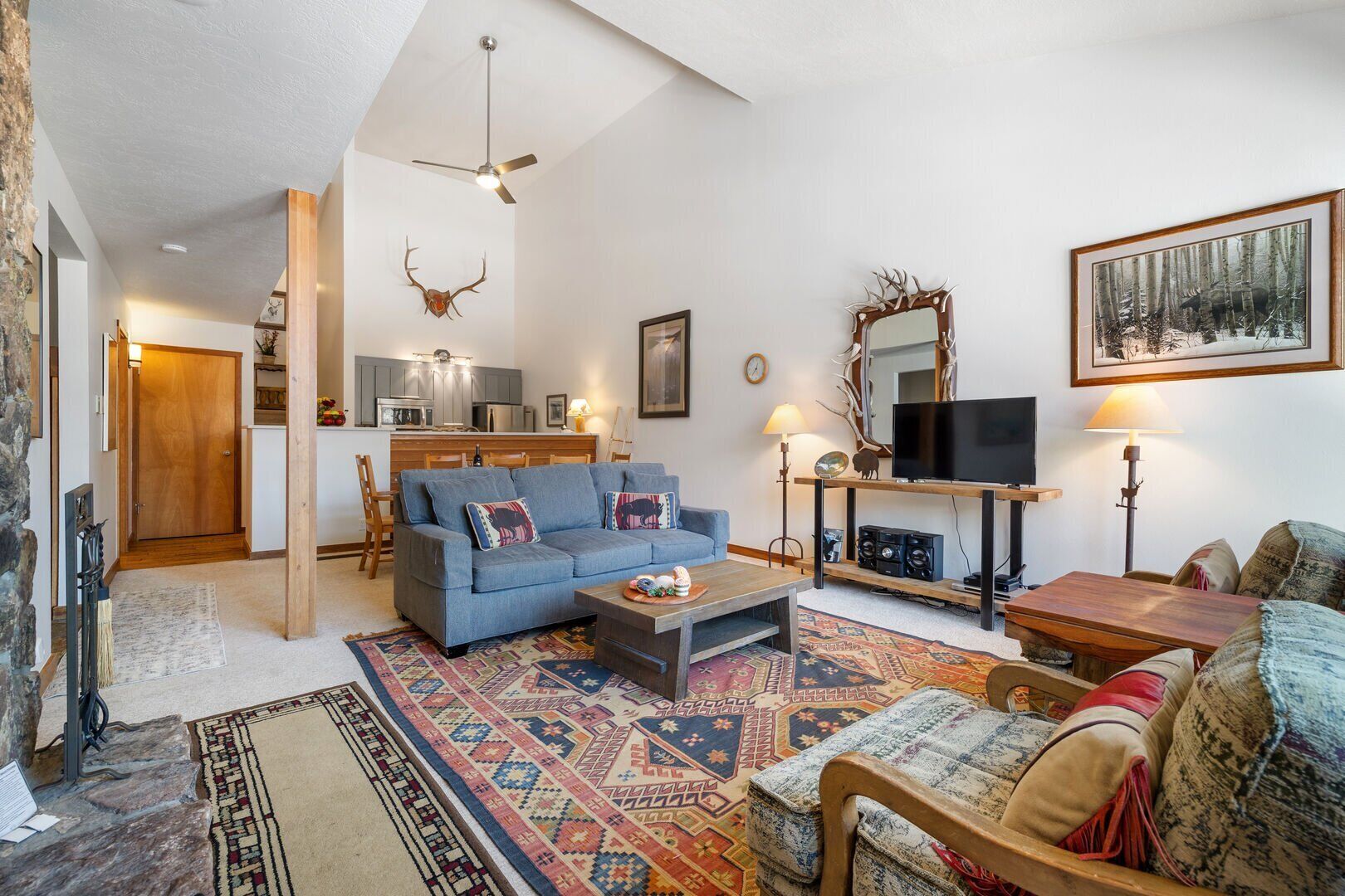 Un salon avec un canapé bleu, une table basse, un téléviseur et une cheminée. Au fond, on aperçoit une cuisine avec un coin repas.