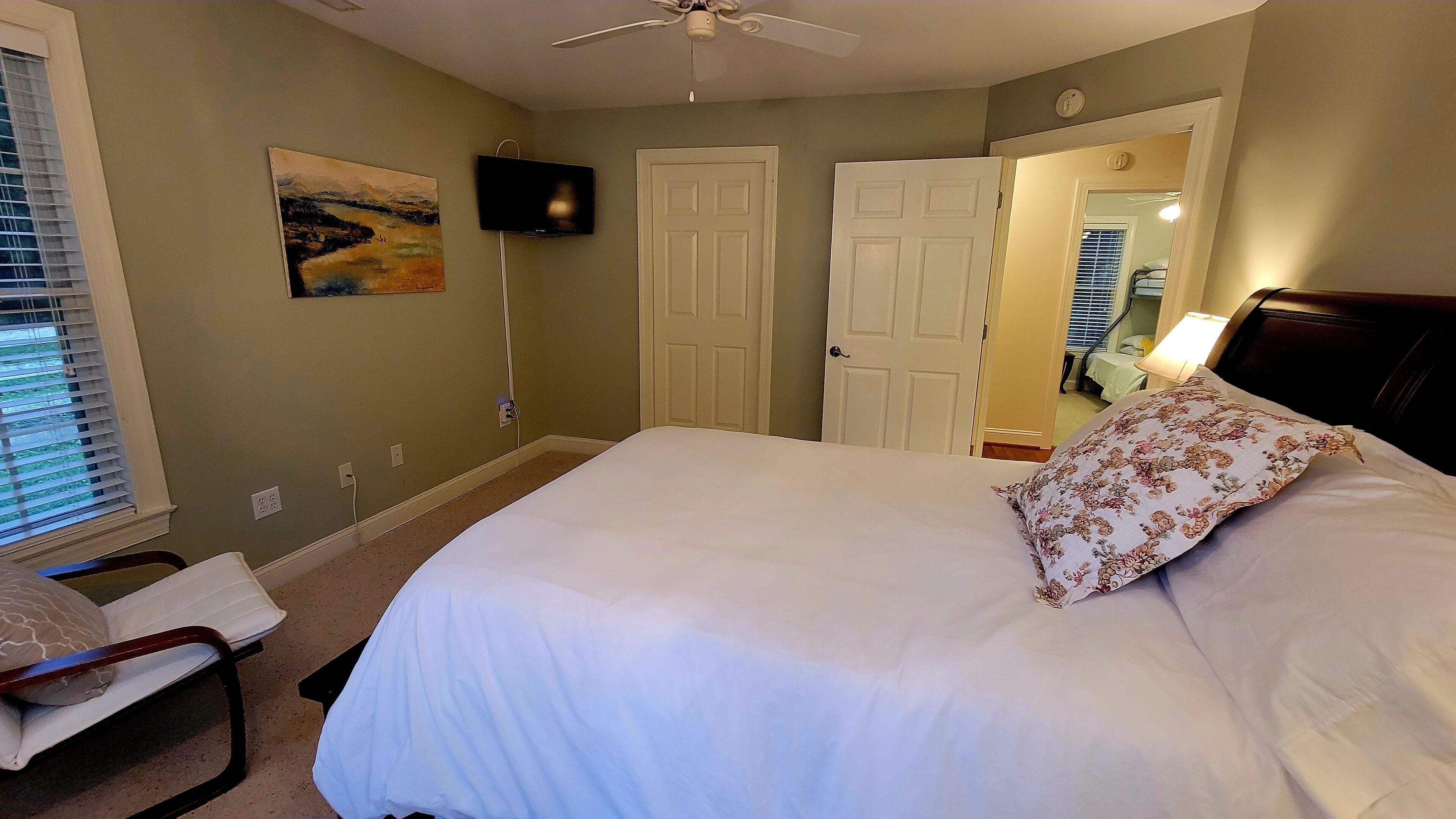 A bedroom with a bed, a chair, a painting, a television, and a door.