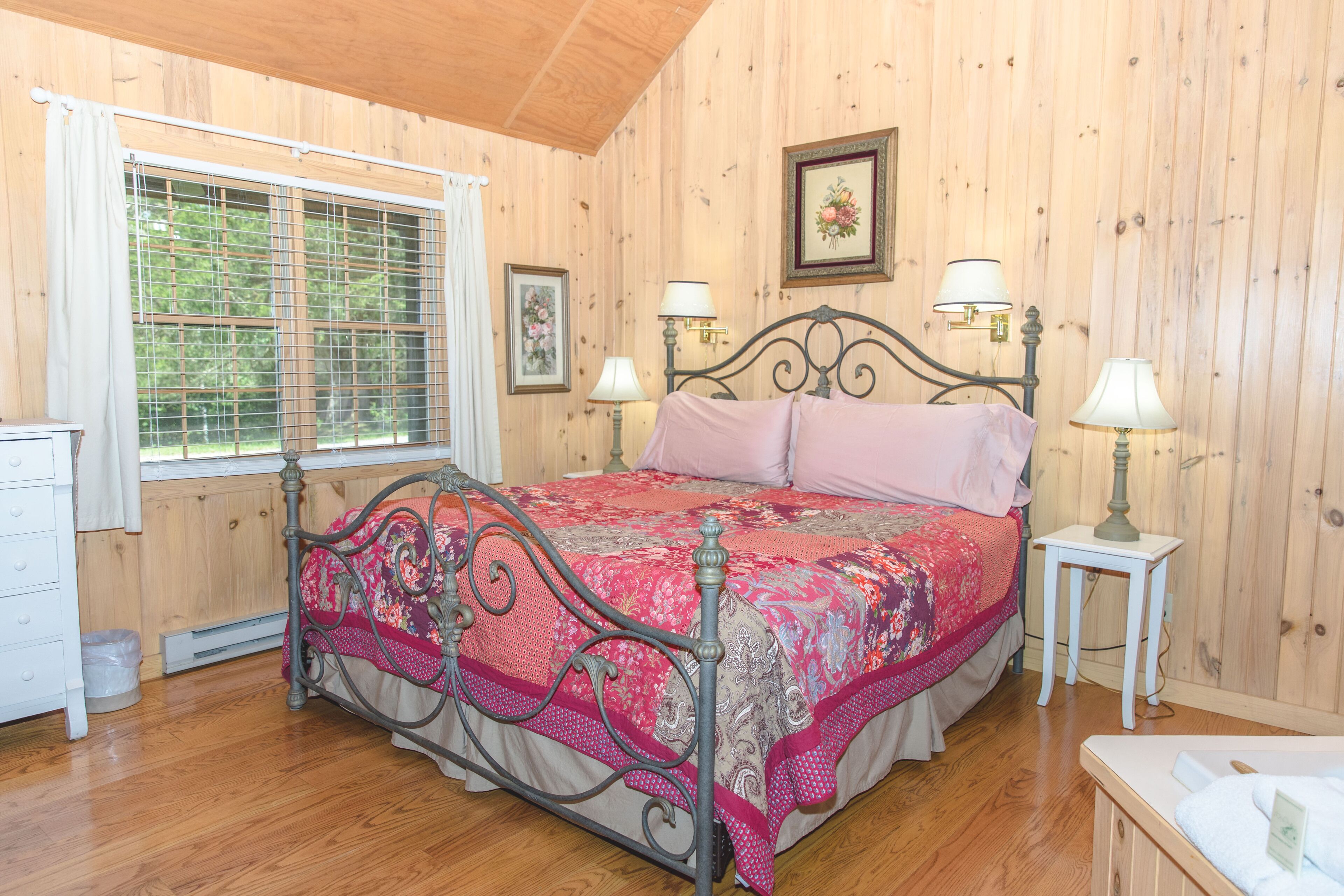 A bedroom with a wooden paneled wall, a metal bed frame, a pink bedspread, two bedside lamps, and a window with a curtain.
