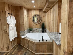A wooden interior with a large bathtub, two white robes, a round mirror, and a potted plant. - Autumn Ridge Cottages (Lexington)