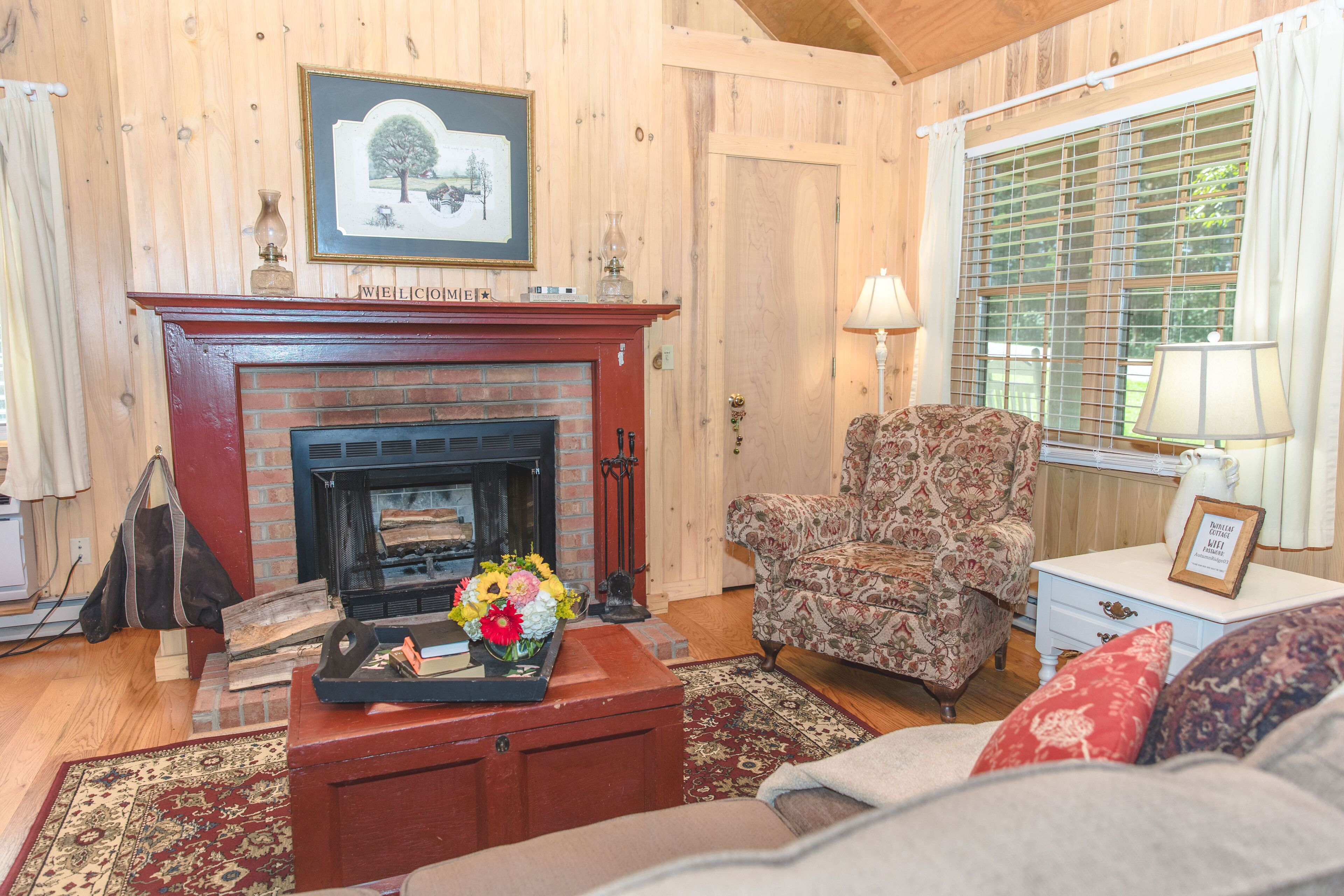 A cozy living room with a fireplace, a patterned armchair, a coffee table with flowers, and a window with blinds.