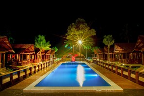 Un área de piscina nocturna con edificios de madera y terraza, un tobogán central y un cielo estrellado.