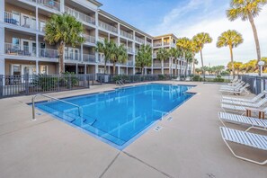 Ein Swimmingpool mit Metallgeländer, umgeben von Liegestühlen und Palmen, mit einem mehrstöckigen Gebäude im Hintergrund.