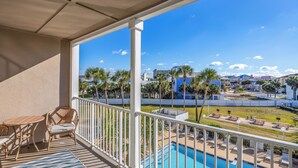 Varanda com móveis de vime, piscina e vista para área de resort com palmeiras e edifícios.