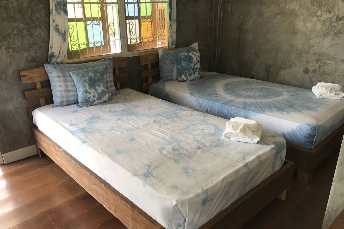 A room with two wooden beds, each with a blue and white patterned bedspread and matching pillows. There is a window with decorative iron bars and a curtain.