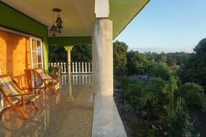 Un balcon abrité offrant une vue sur un paysage luxuriant, un coin salon avec des chaises à rayures et une balustrade.