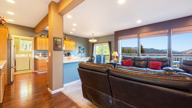 A modern kitchen with wooden cabinets, a dining area with a leather couch, and a large window offering a scenic view. - West Sail - Play & Work in Hood River! River & Mountain Views, Quiet, Walk to Downtown (Hood River)