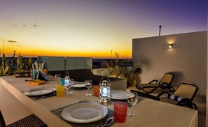 Mesa de jantar posta para dois com vista para o pôr do sol, um copo de coquetel e uma garrafa de água.