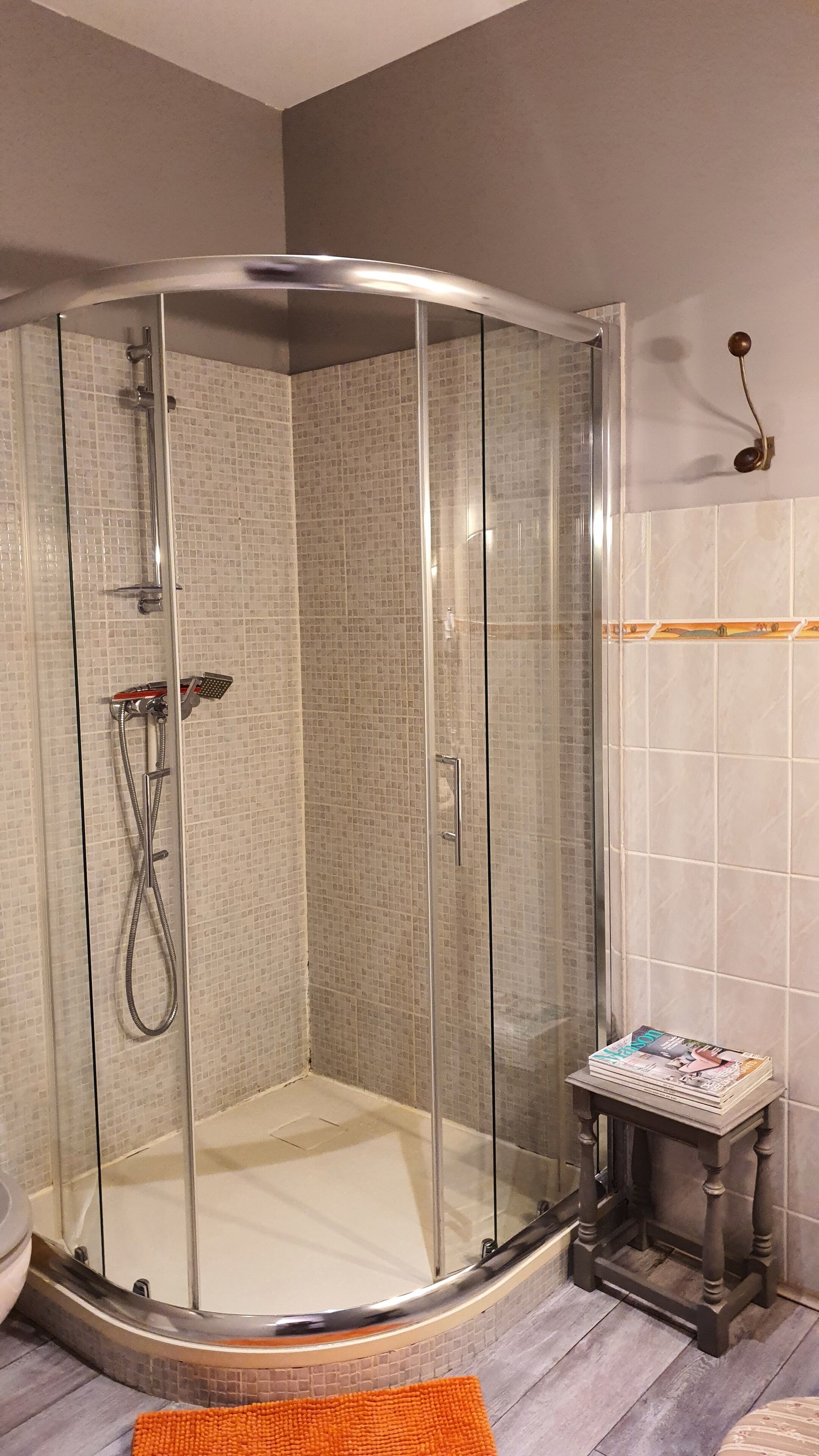 Eine gläserne Dusche mit gefliesten Wänden, ein kleiner Holzschemel mit Zeitschriften und ein Regal mit einem dekorativen Gegenstand.