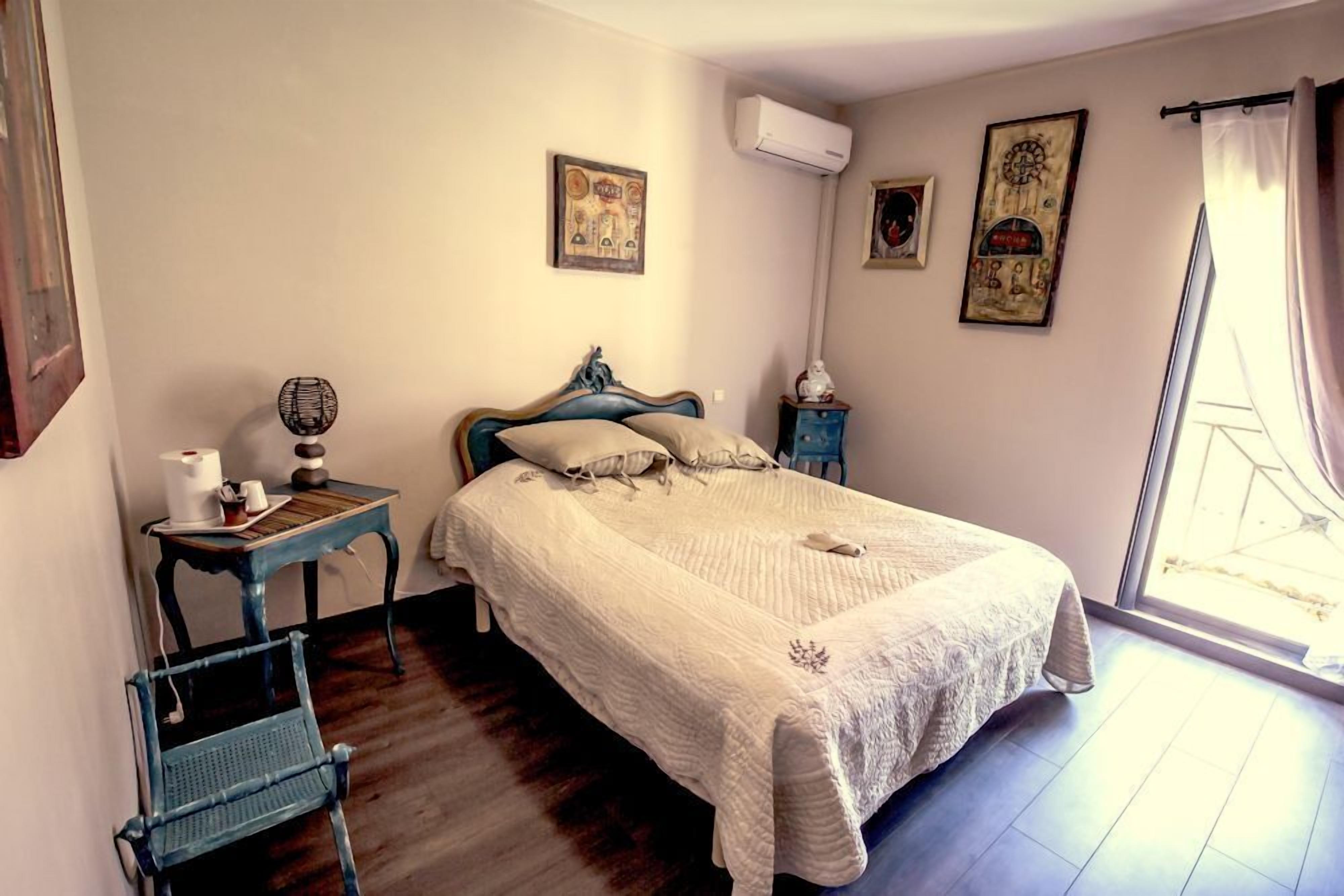 Een slaapkamer met een bed, een tafeltje met een lamp, een stoel en een raam met gordijnen.
