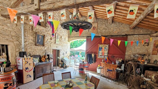 A rustic dining area with a large table, wooden chairs, and a stone wall backdrop. - Le Clos des Aramons (Bernis)
